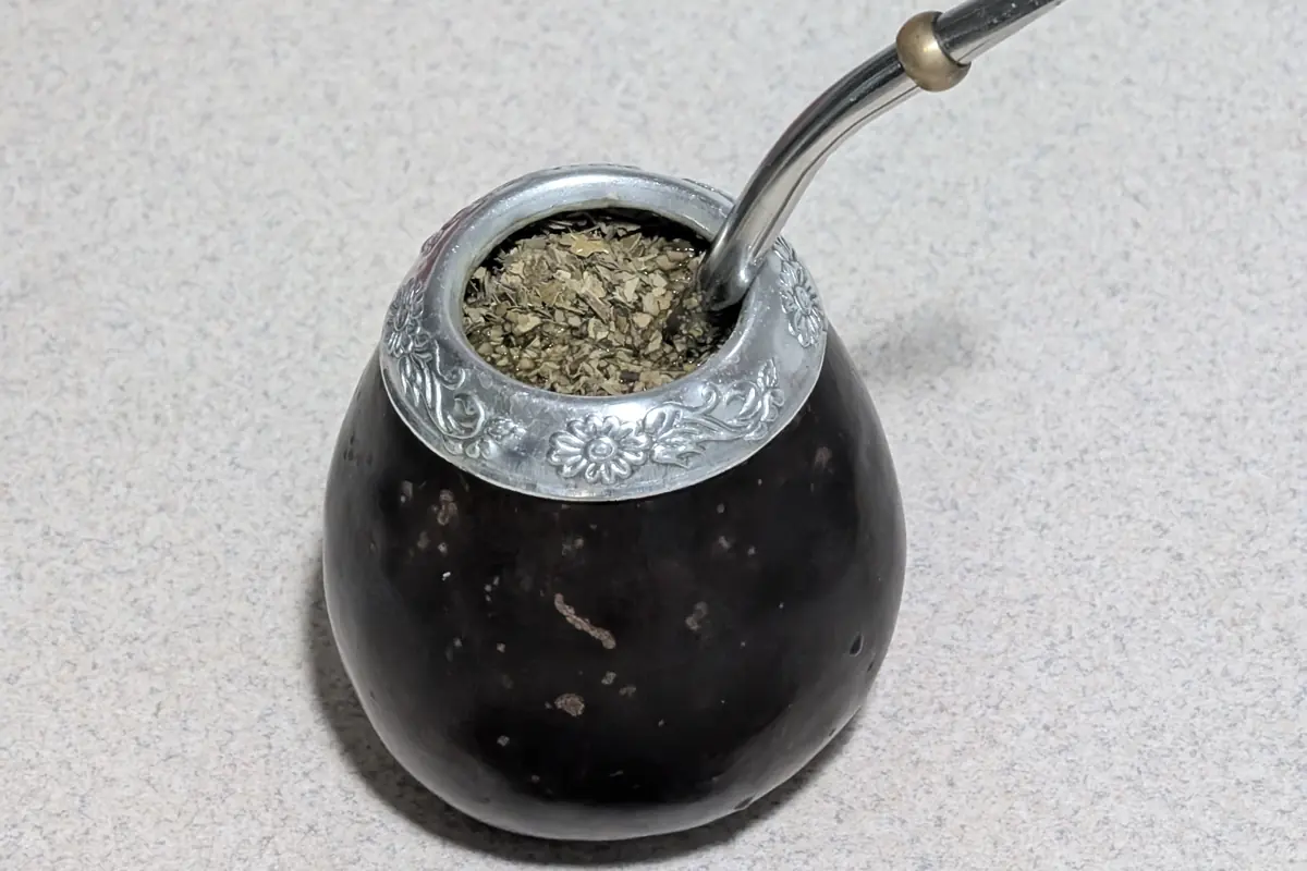 A black mate gourd with a silver rim, filled with yerba mate and a bent metal bombilla straw, beside a wooden scoop
