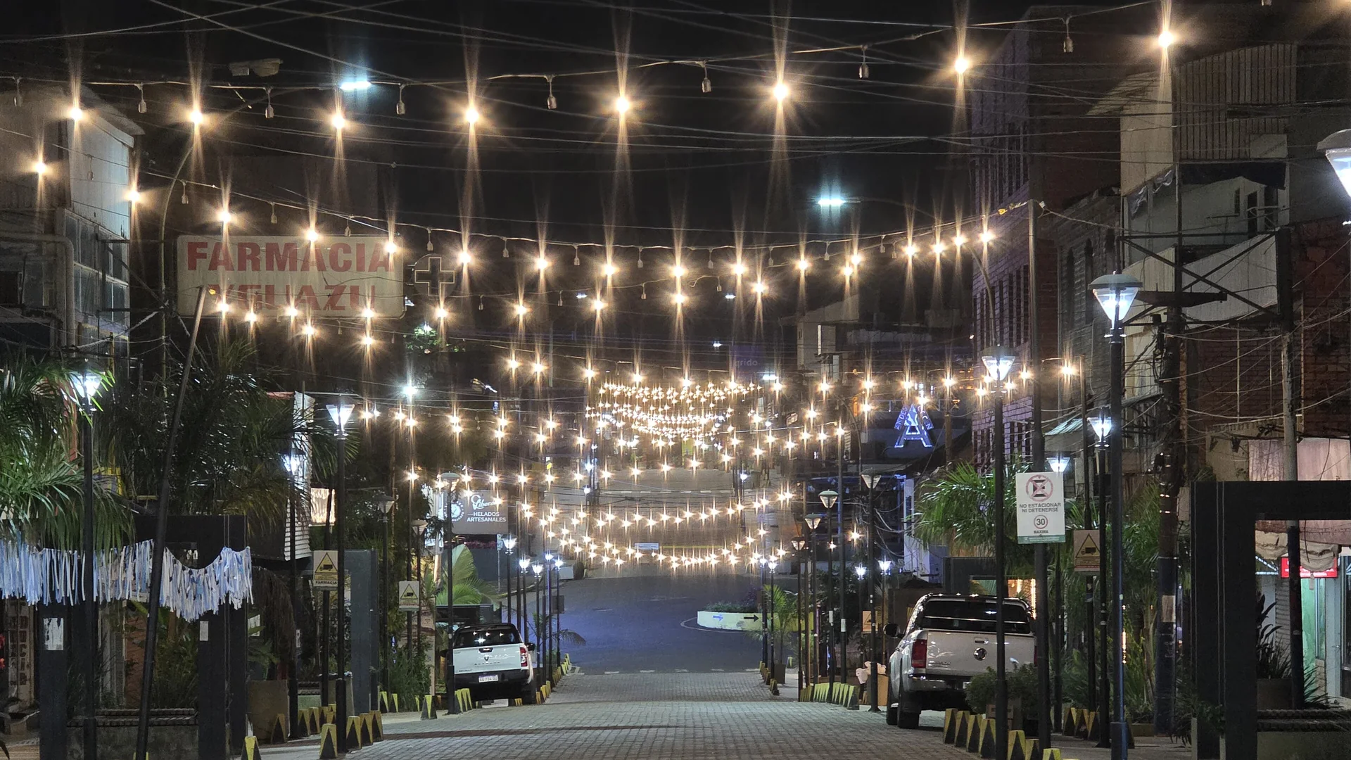 Avenida Brasil in Puerto Iguazú at night, a cobbled street under strings of lights, with palms and shopfronts on both sides.